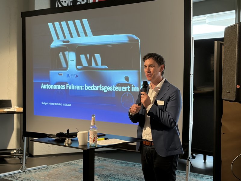 Foto: Ein Mann im Anzug hält einen Vortrag. Neben ihm auf einer Leinwand sieht man die Titelfolie seiner Präsentation