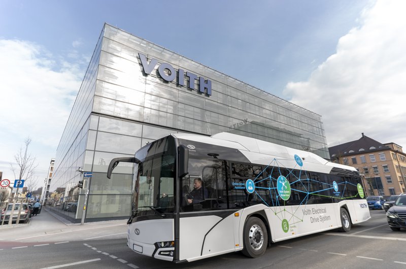 Ein weißer Bus vor einem verglasten Firmengebäude, an dem Voith steht