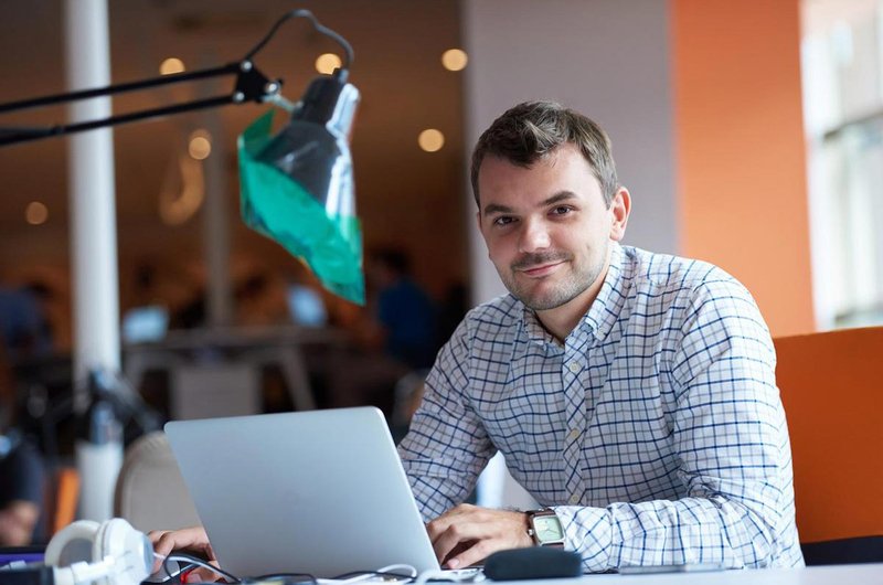 Ein junger Mann im Hemd sitzt am Schreibtisch auf dem ein Laptop steht.