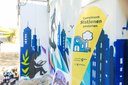 Gelber Aufsteller mit der Aufschrift 'Gemeinsam Stationen gestalten' vor einer Wand mit buntem Graffiti, das eine Stadtlandschaft und eine sitzende Person zeigt