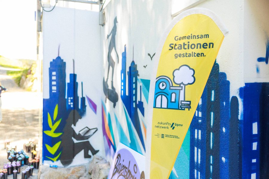 Gelber Aufsteller mit der Aufschrift 'Gemeinsam Stationen gestalten' vor einer Wand mit buntem Graffiti, das eine Stadtlandschaft und eine sitzende Person zeigt