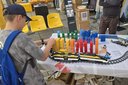 Junge mit blauer Kappe und Rucksack baut an einem Modell aus bunten Bausteinen auf einem Tisch mit Schienen und Zügen