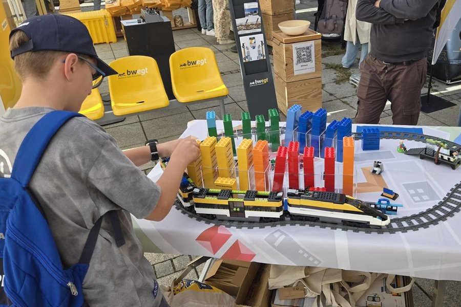 Junge mit blauer Kappe und Rucksack baut an einem Modell aus bunten Bausteinen auf einem Tisch mit Schienen und Zügen