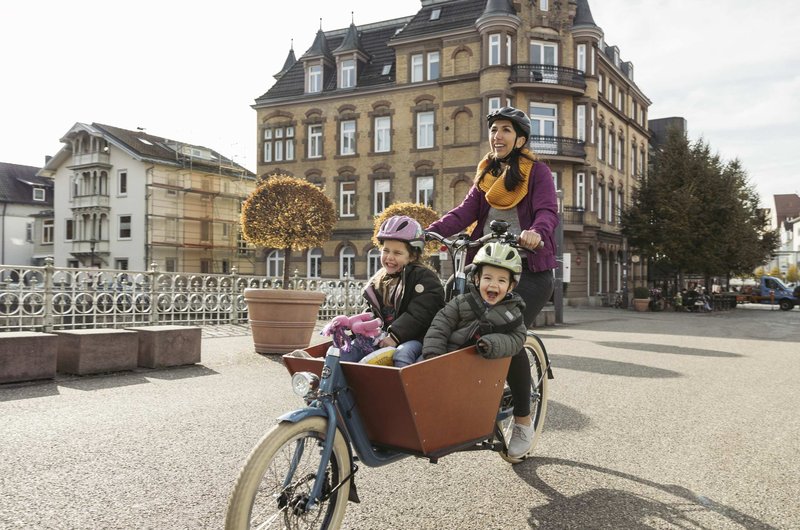 Lastenrad mit zwei Kindern in der Transportbox auf einer Brücke in einer Innenstadtumgebung.