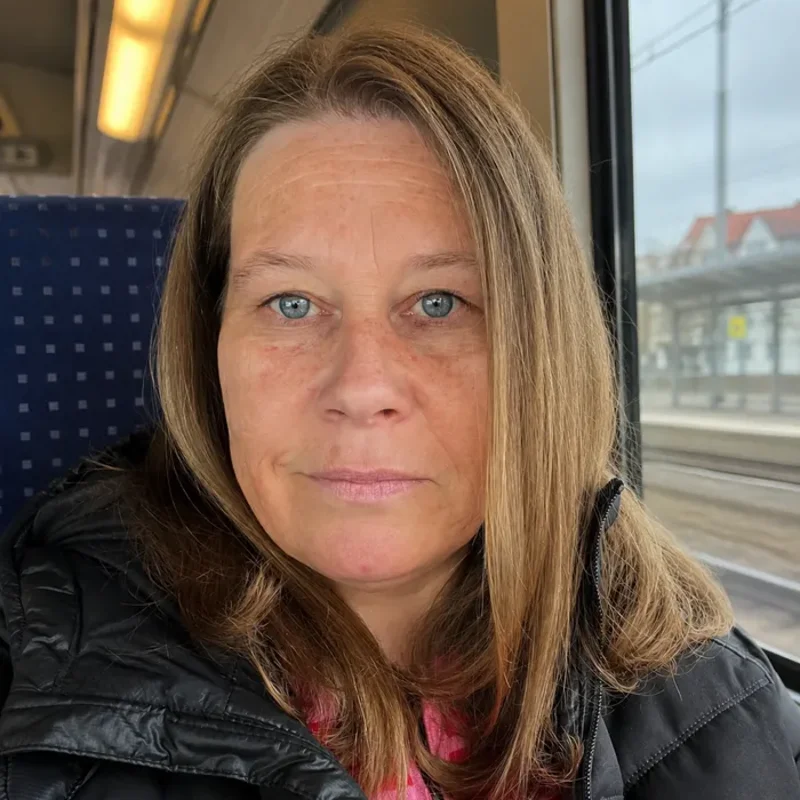Frau mit schulterlangen blonden Haaren und blauen Augen sitzt in einem Zugabteil und trägt eine schwarze Jacke