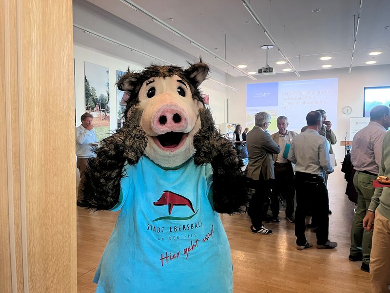 Person im Wildschwein-Kostüm mit blauem T-Shirt und Aufdruck 'Stadt Ebersbach Hier geht was' vor einer Gruppe von Menschen in einem hellen Raum