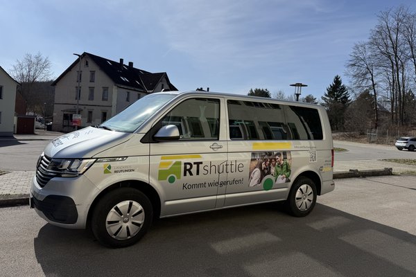 Ein weißer Kleinbus des Angebots RTshuttle des Landkreises Reutlingen steht auf einem Parkplatz. Auf der Fahrzeugseite sind das RTshuttle-Logo, der Schriftzug Kommt wie gerufen! sowie ein Bildmotiv und ein QR-Code zu sehen. Im Hintergrund befinden sich Gebäude, Bäume und ein heller Himmel.