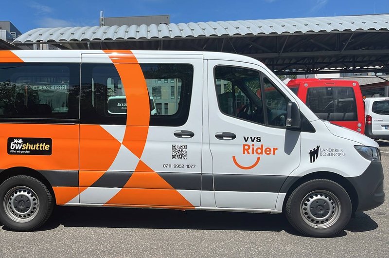 Weißer Kleinbus mit orangefarbenem Design und den Aufschriften 'VVS Rider' und 'ÖV shuttle' auf einem Parkplatz