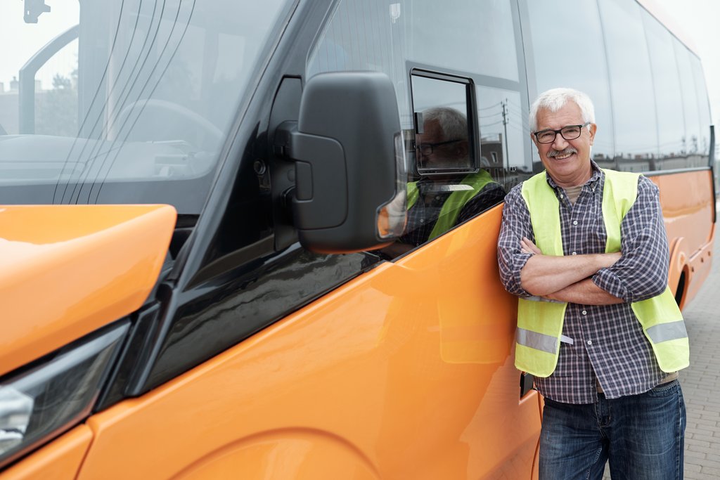 Ein älterer Busfahrer steht an einen orangenen Bus angelehnt. Man sieht den Mann aus einer schrägen Seitenperspektive. Er hat die Arme verschränkt, trägt eine Gelbe Warnweste und lächelt in die Kamera.