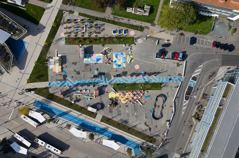 Senkrechte Luftaufnahme einer temporär umgestalteten Platzfläche mit bunten Bodenillustrationen, Sitz- und Spielbereichen, Pflanzinseln und Mobilitätsflächen. Die Fläche ist von Straßen, Gebäuden und öffentlichen Verkehrsflächen umgeben.