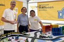 Drei Personen stehen an einem Tisch mit einer Modelleisenbahn und bunten Legosteinen vor gelben Bannern mit Texten zu Bahnthemen.