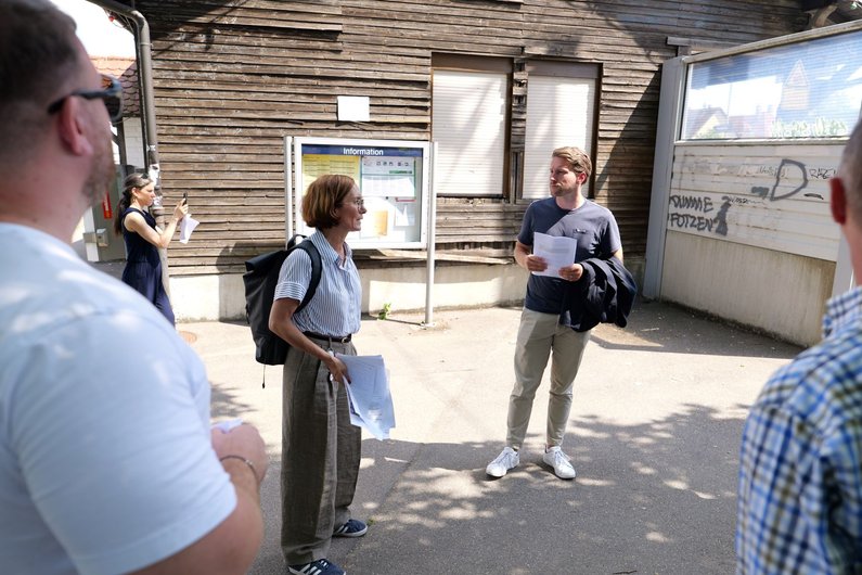 Gruppe von Menschen steht im Freien vor einem hölzernen Gebäude und hält Papiere in der Hand