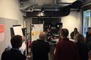 Gruppe von Menschen steht in einem Raum und hört einem Mann zu, der vor einem Flipchart steht und spricht