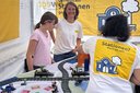 Drei Personen an einem Tisch mit Modellbahngleisen und kleinen Gebäuden, eine trägt ein weißes T-Shirt mit gelbem Aufdruck und einer stilisierten blauen Lokomotive