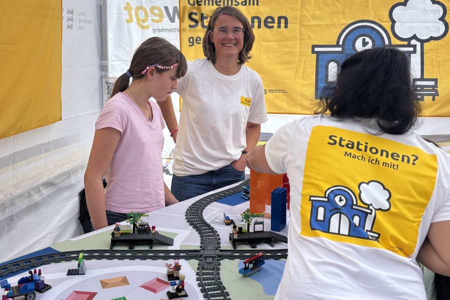 Drei Personen an einem Tisch mit Modellbahngleisen und kleinen Gebäuden, eine trägt ein weißes T-Shirt mit gelbem Aufdruck und einer stilisierten blauen Lokomotive