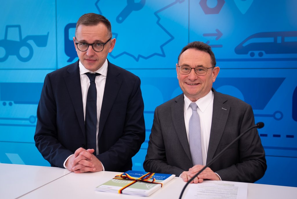 Zwei Männer in Anzügen sitzen an einem Tisch und unterzeichnen Dokumente vor einer blauen Wand.