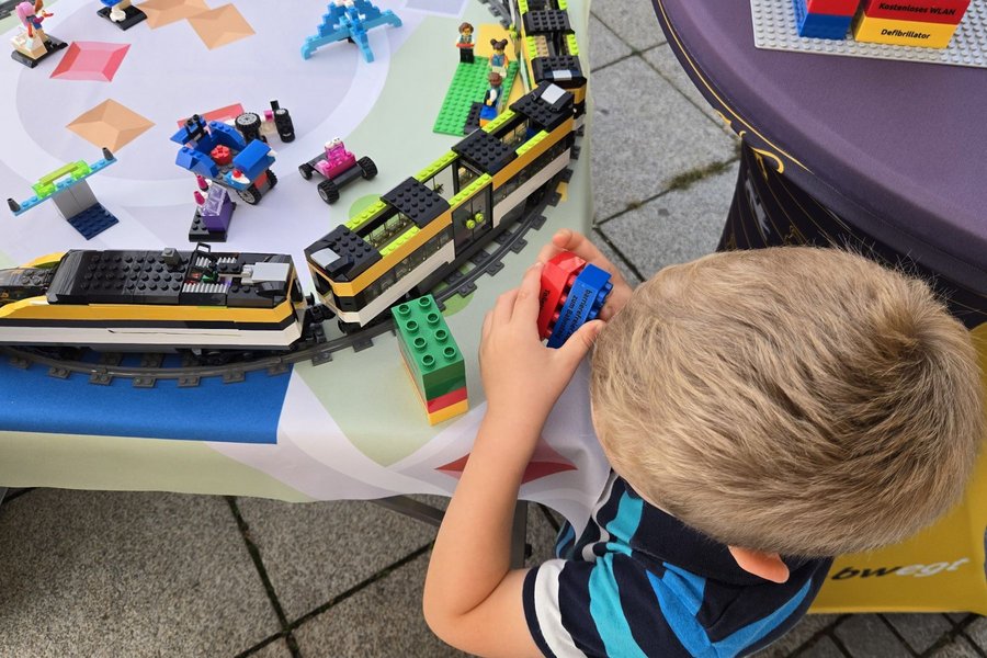 Kind baut mit bunten LEGO-Steinen an einem Modellzug auf einem Tisch mit Schienen und weiteren LEGO-Fahrzeugen