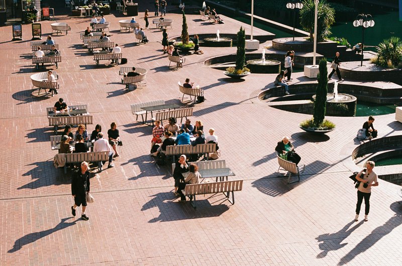 Menschen sitzen und bewegen sich auf einem großzügigen öffentlichen Platz mit Bänken, Tischen und Springbrunnen.