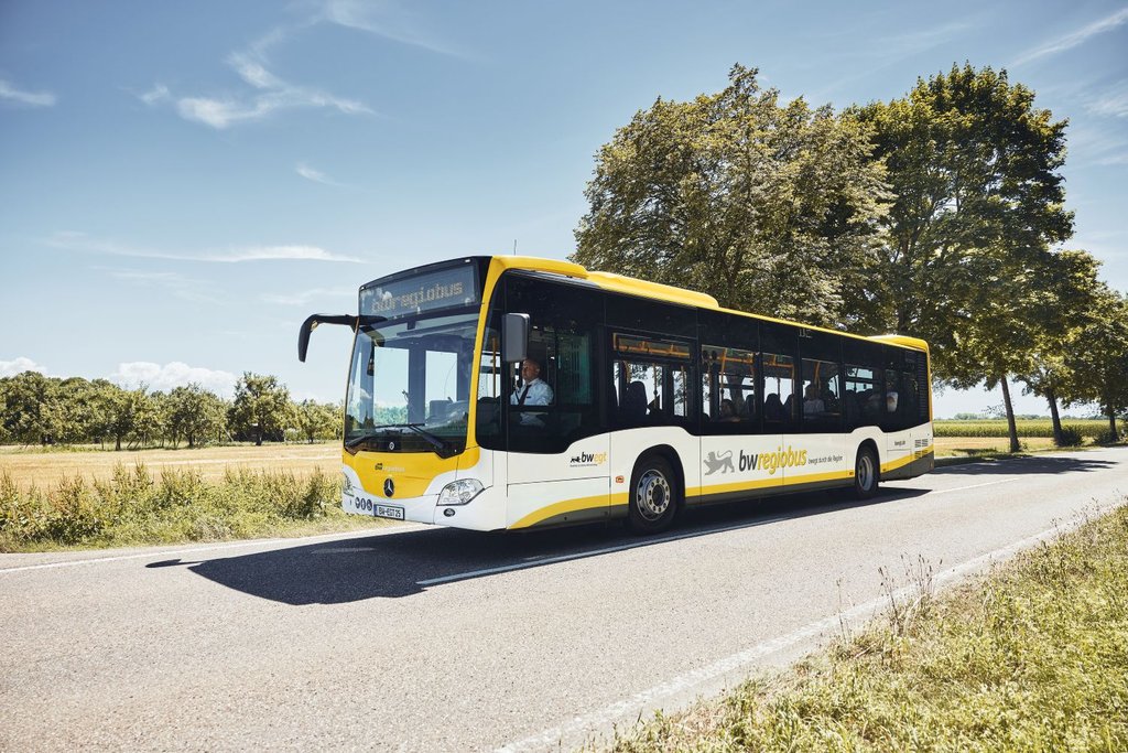 Ein Regiobus im gelb-weißen Landesdesign fährt auf einer Landstraße. Im Hintergrund sind abgeerntete Felder und Bäume zu sehen.