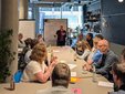 Mehrere Menschen sitzen um einen langen Tisch. Vorne steht eine Frau vor einem Flipchart und erklärt etwas.