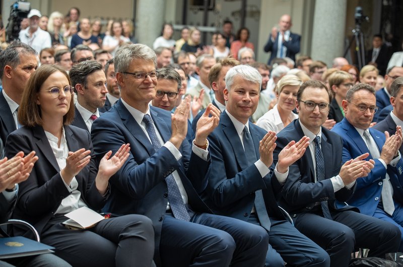 Im Vordergrund sitzen mehrere Personen in dunklen Anzügen und hellen Hemden nebeneinander und applaudieren. Die meisten tragen Brillen und wirken konzentriert. Im Hintergrund sind zahlreiche weitere Menschen zu sehen, die ebenfalls aufmerksam nach vorne blicken. Die Szene spielt in einem großen, hellen Raum mit hohen Säulen und Tageslicht. Die Atmosphäre wirkt formell und feierlich.