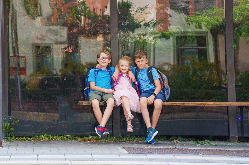Drei blonde Schulkinder sitzen auf einer Bank an einer Bushaltestelle.