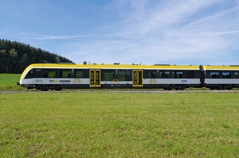 Ein weiß-gelber Regionalzug im bwegt-Design fährt durch eine Wiesenlandschaft.