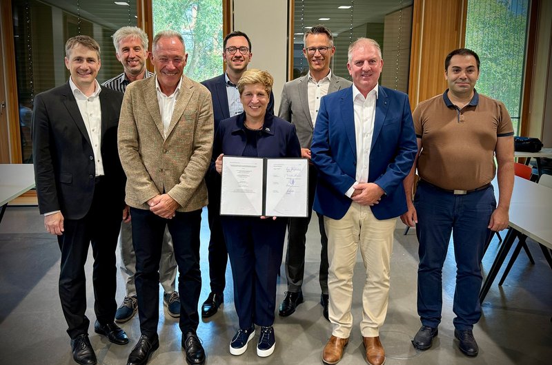 Gruppenfoto mit acht Personen in einem Raum; die Ministerin Razavi steht in der Mitte und hält ein geöffnetes Dokument mit Unterschriften in den Händen.
