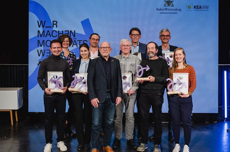 Baden-Württembergs Verkehrsminister Winfried Hermann steht gemeinsam mit den Gewinnerinnen und Gewinnern der Landesauszeichnung "Wir machen Mobilitätswende" auf einer Bühne. 