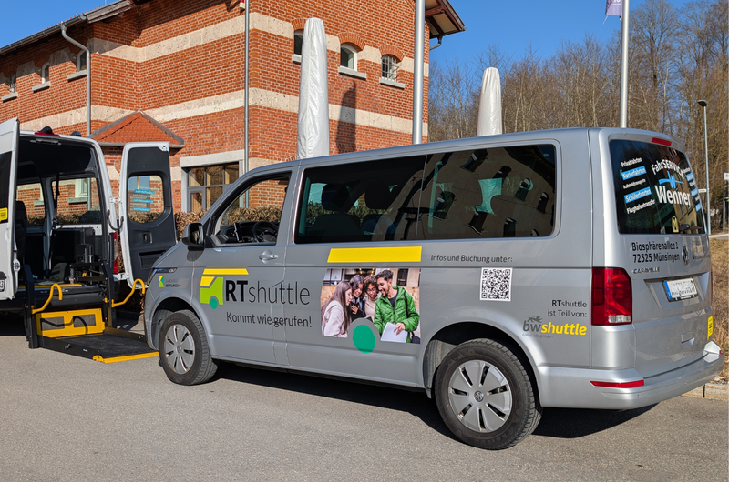 Ein silberner Kleinbus, das RTshuttle, unter anderem beklebt mit dem Logo, dem Slogan Kommt wie gerufen! sowie dem Logo bwshuttle des Landes Baden-Württemberg, steht am Straßenrand vor einem großen Backsteingebäude. Vor dem Kleinbus steht ein weiteres wartendes Fahrzeug.