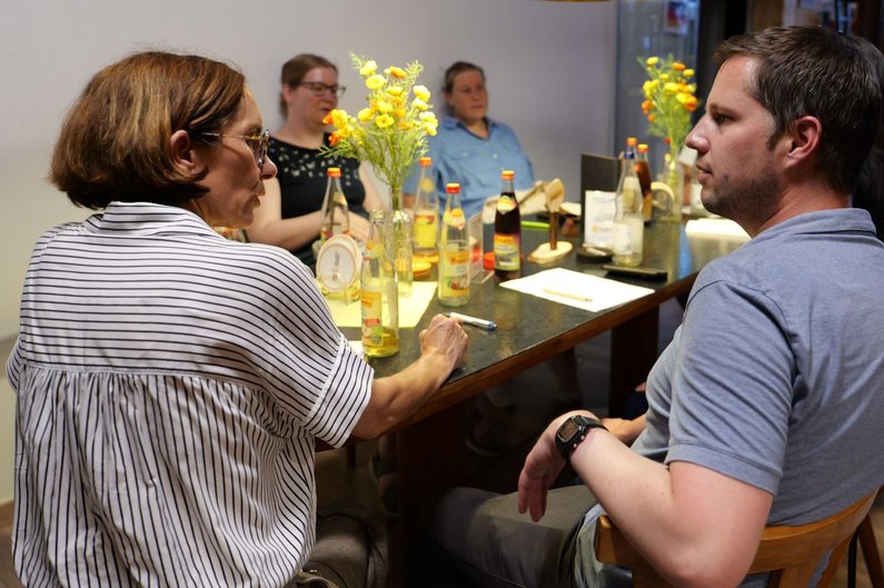 Vier Personen sitzen an einem Tisch mit mehreren Glasflaschen und zwei Vasen mit gelben Blumen in einem Innenraum