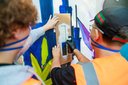Zwei Personen mit Schutzbrillen und Warnwesten sprühen mit Schablone ein Graffiti an eine Wand