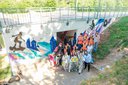 Gruppe von Jugendlichen mit Maleranzügen und orangenen Westen vor einer farbenfrohen Wandbemalung unter einer Brücke mit Skateboard- und Fahrradmotiven.