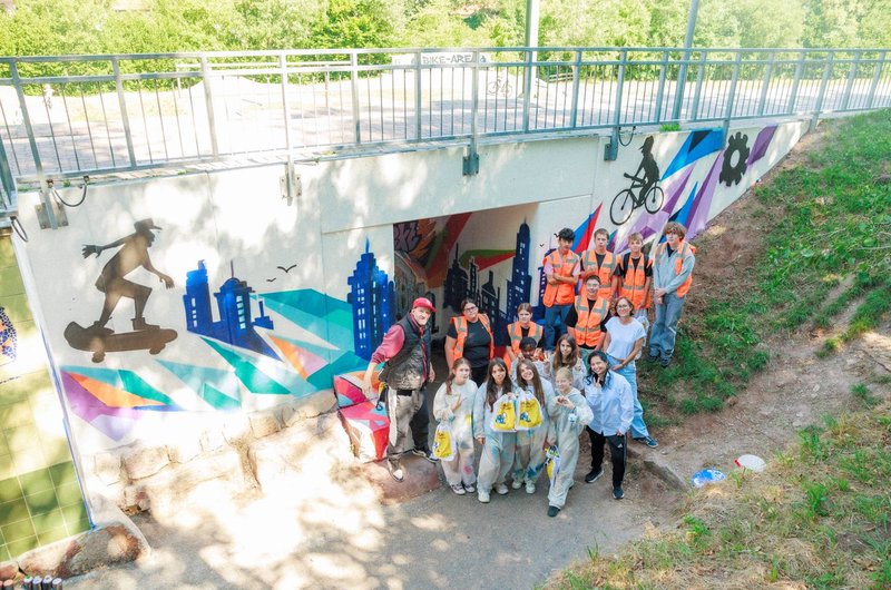 Gruppe von Jugendlichen mit Maleranzügen und orangenen Westen vor einer farbenfrohen Wandbemalung unter einer Brücke mit Skateboard- und Fahrradmotiven