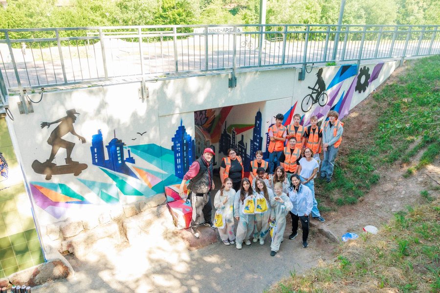 Gruppe von Jugendlichen mit Maleranzügen und orangenen Westen vor einer farbenfrohen Wandbemalung unter einer Brücke mit Skateboard- und Fahrradmotiven.