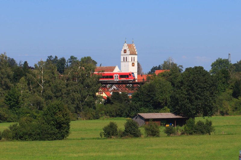 Ein roter Regionalzug fährt auf einer Brücke vor einem weißen kirchturm vorbei. Rechts und link stehen Bäume, im Vordergrund ist eine Wiese zu sehen.