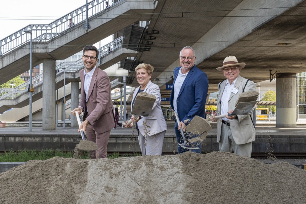 Vier Personen in formeller Kleidung stehen mit Schaufeln vor einem großen Sandhaufen unter einer Brücke an einem Bahnhof