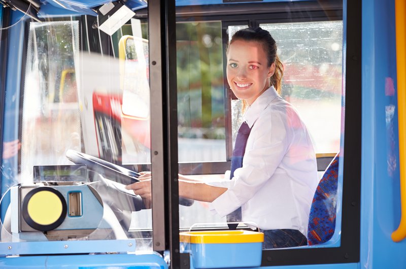 Eine Busfahrerin in einem weißen Hemd sitzt am Steuer eines blauen Busses.