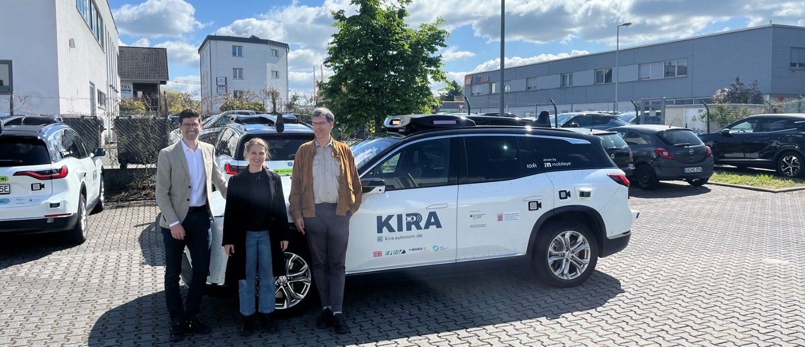 Drei Personen stehen vor einem weißen SUV mit der Aufschrift 'KIRA' auf einem gepflasterten Parkplatz vor Industriegebäuden bei sonnigem Himmel
