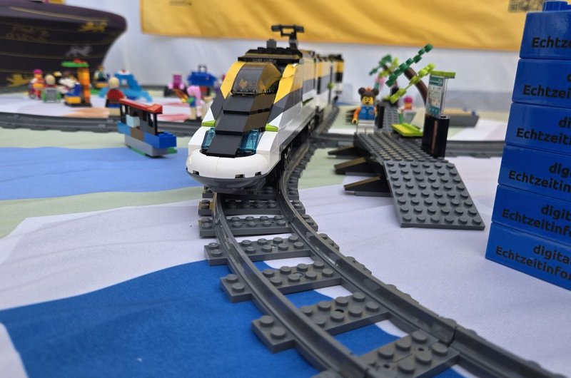 Nahaufnahme eines weißen und schwarzen LEGO-Zuges auf grauen Schienen vor einem gelben Banner mit blauem Text