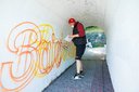 Person mit roter Mütze, schwarzer Weste und Shorts sprüht bunte Schrift an die Wand eines weißen Tunnels