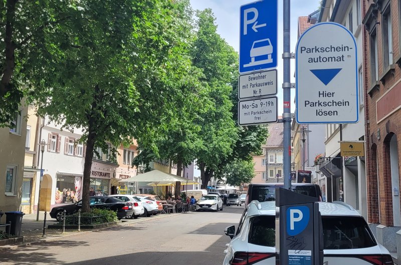 Eine Straße in der ein Auto parkt, aber auch viel grün ist und ein Café den Parkraum als Sitzbereich nutzt.
