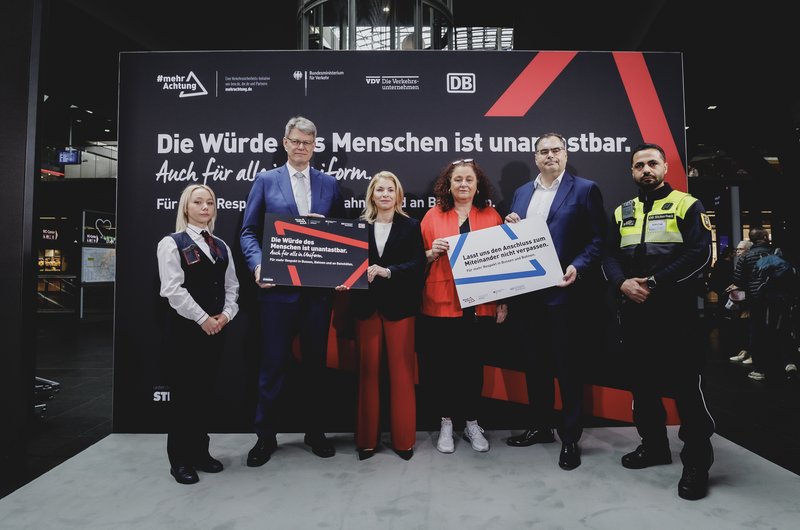 Sechs Personen stehen vor einer großen schwarzen Wand mit weißer und roter Schrift, zwei halten Schilder mit Texten zur Würde des Menschen und Respekt, eine Person trägt eine Uniform der DB-Sicherheit.