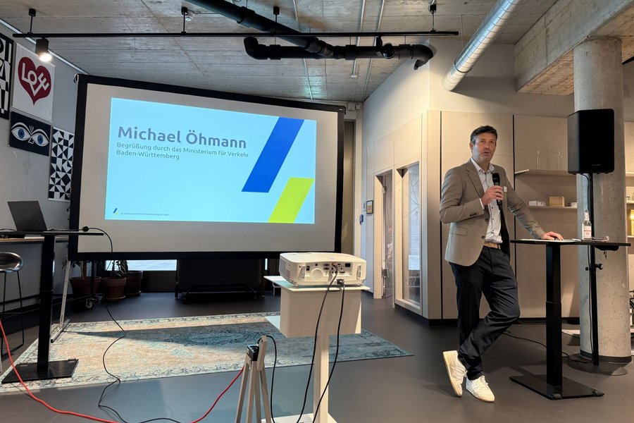 Mann mit Mikrofon lehnt an Stehtisch vor Leinwand mit Präsentationsfolie 'Michael Öhmann' in modernem Raum mit sichtbaren Rohren an der Decke