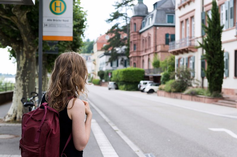 Eine junge Frau mit einem roten Rucksack steht in Heidelberg an einer Bushaltestelle und wartet auf den Bus.