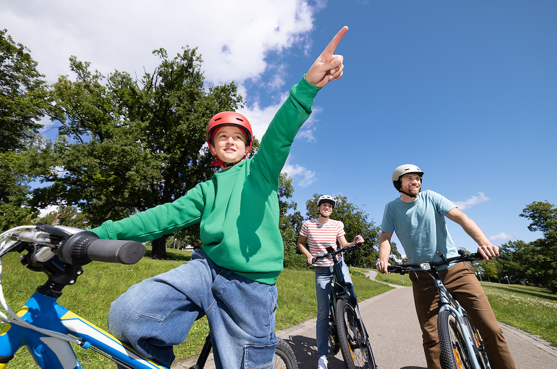 Kind mit rotem Fahrradhelm auf einem Fahrrad zeigt im Vordergrund mit dem Finger nach vorne, dahinter zwei erwachsene Personen auf Fahrrädern auf einem Radweg im Grünen.