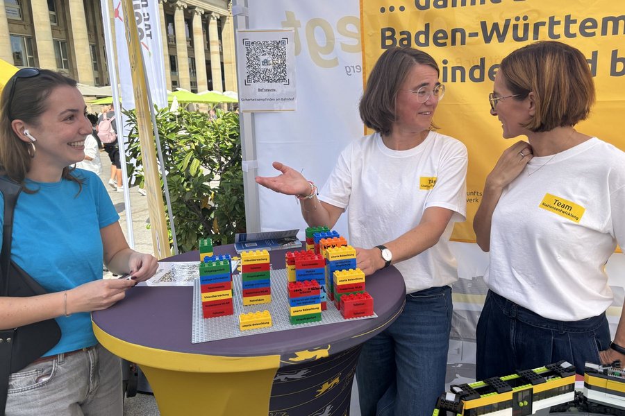 Drei Frauen unterhalten sich an einem Stehtisch mit bunten Lego-Steinen vor einem gelben Banner mit Textfragmenten zu Baden-WürttembergDrei Frauen unterhalten sich an einem Stehtisch mit bunten Lego-Steinen vor einem gelben Banner mit Textfragmenten zu Baden-Württemberg