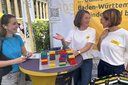 Drei Frauen unterhalten sich an einem Stehtisch mit bunten Lego-Steinen vor einem gelben Banner mit Textfragmenten zu Baden-WürttembergDrei Frauen unterhalten sich an einem Stehtisch mit bunten Lego-Steinen vor einem gelben Banner mit Textfragmenten zu Baden-Württemberg