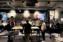 Gruppe von acht Personen steht um zwei nebeneinanderstehende Tische in einem modernen, dunklen Raum und betrachtet Papiere auf den Tischen
