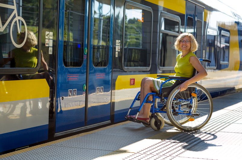 Eine Frau im Rollstuhl, ist an einer blauen Tür eines gelb, weiß, blauen Zuges.  